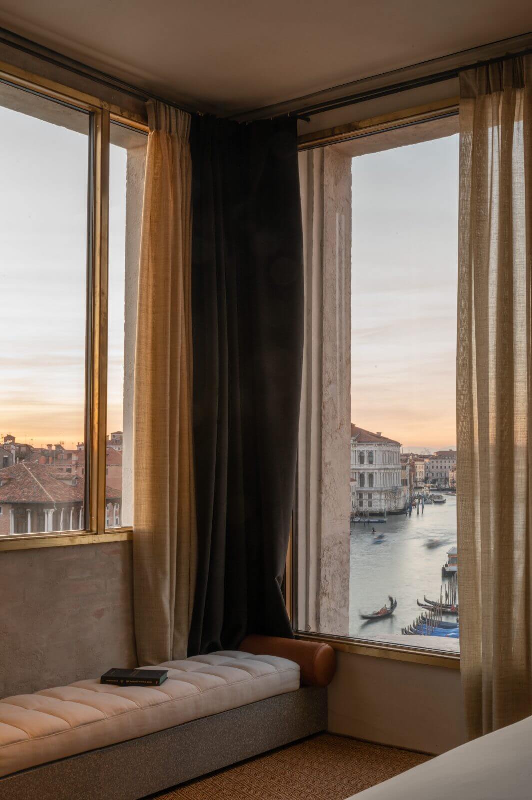 The Venice Venice Hotel, Venice, Italy — Corner window seat overlooking Grand Canal at sunset, brass-framed windows with layered linen curtains, upholstered bench, Venetian architecture and gondolas visible across water.