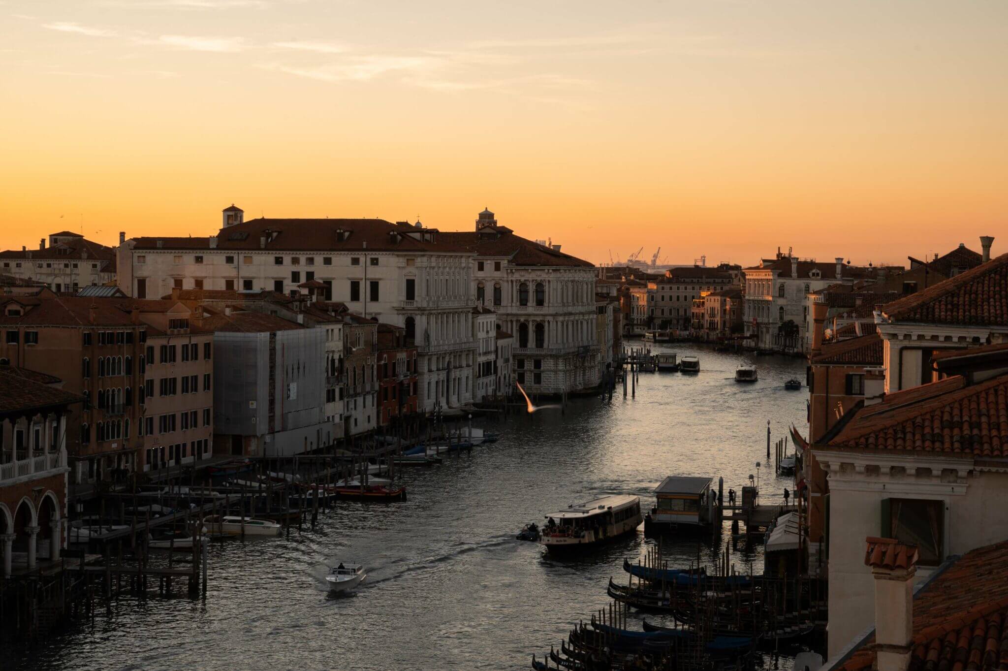 The Venice Venice Hotel, Venice, Italy — Golden hour light illuminates Venetian palazzo architecture along the Grand Canal with moored boats and historic waterfront buildings in warm ochre and cream tones.