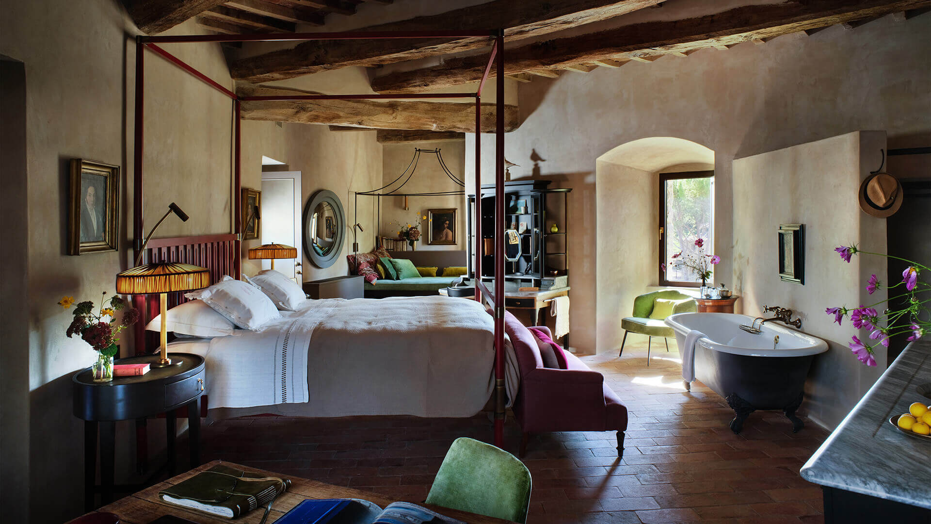 Castello di Reschio, Europe — Rustic bedroom with canopy bed, exposed wooden beams, arched stone doorway, freestanding dark bathtub, and terracotta floors flooded with natural light.
