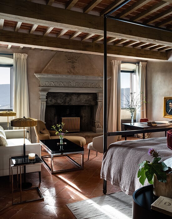 Castello di Reschio, Europe — Canopied bedroom with fireplace, exposed beam ceiling, terracotta floors, natural light, and modern furnishings in historic stone interior.