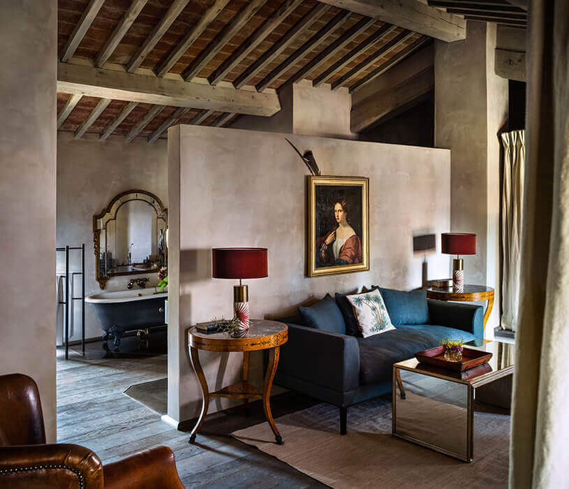 Castello di Reschio, Europe — Rustic bedroom with exposed wooden beams, blue velvet sofa, gilt-framed portrait, marble sink, and warm lamplight in stone-walled suite.