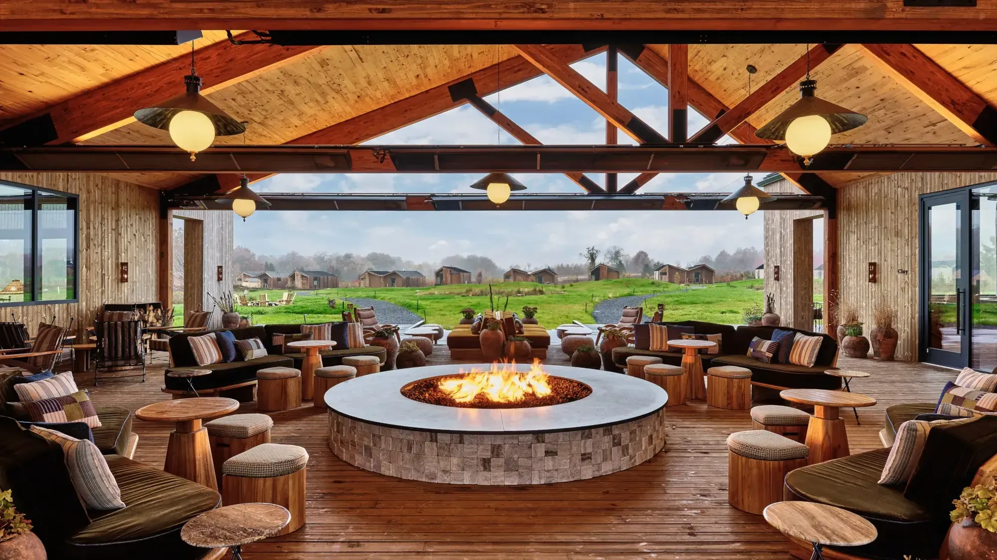 The Great Porch — Great Porch, North America — Open-air pavilion with exposed timber beams, circular fire pit, lounge seating, and pastoral views across manicured grounds.