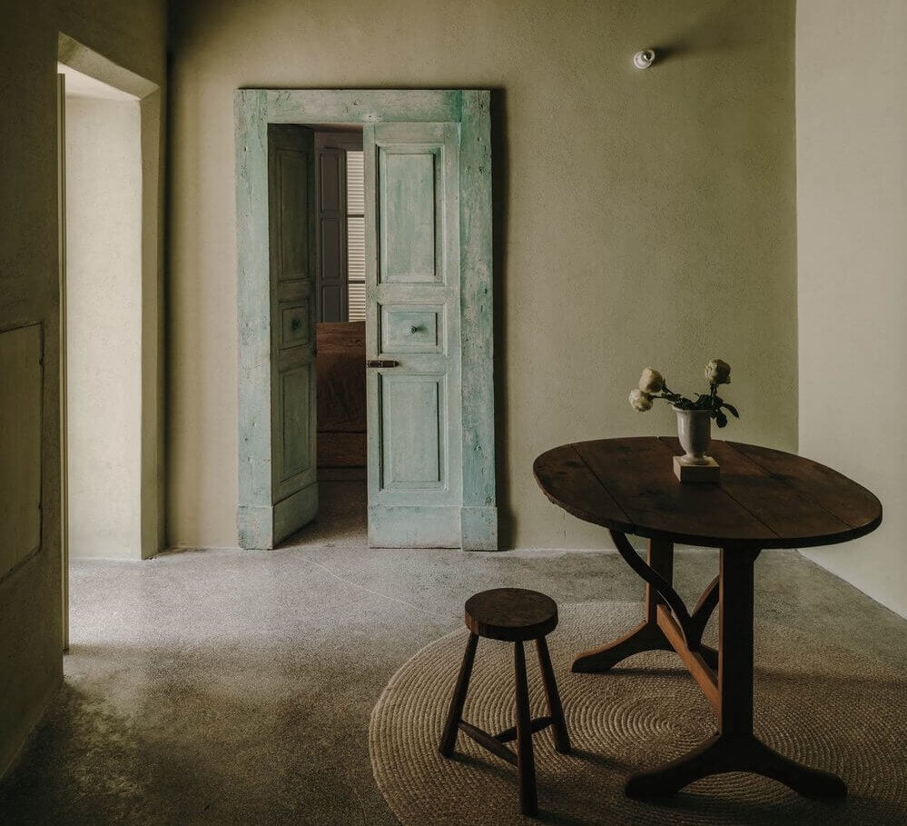 Casa Soleto, Europe — Minimalist room with sage green walls, weathered turquoise doors, dark wood table and stool, natural light, modernist interior design.
