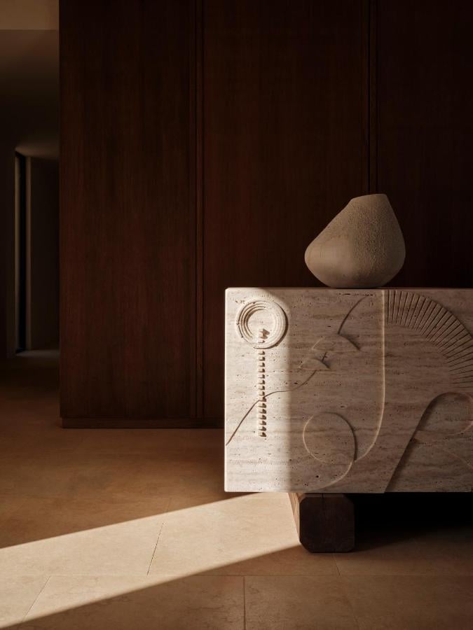 Gundari, Greece — Sculptural wooden console with carved details and travertine vessel, minimalist interior with warm sunlight and dark wood paneling.