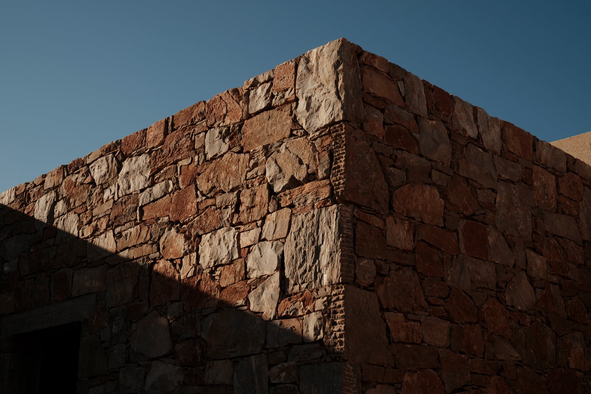 Gundari, Greece — Weathered stone facade with warm terracotta and cream masonry, sharp afternoon shadows, austere architectural detail.