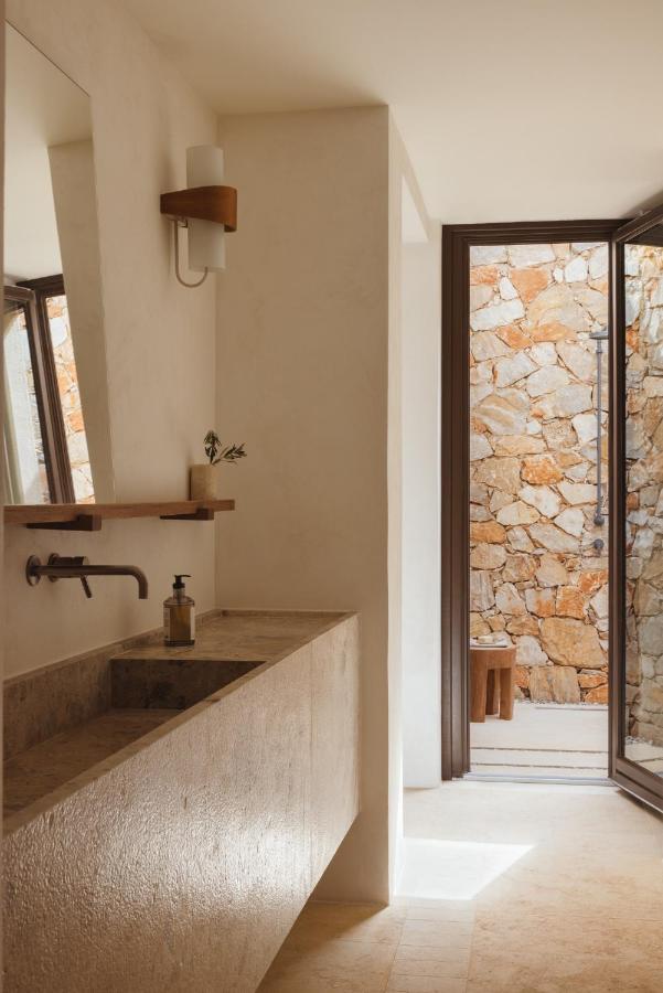 Gundari, Greece — Minimalist bathroom with stone sink, dark fixtures, timber shelving, and open door revealing terracotta stone courtyard.