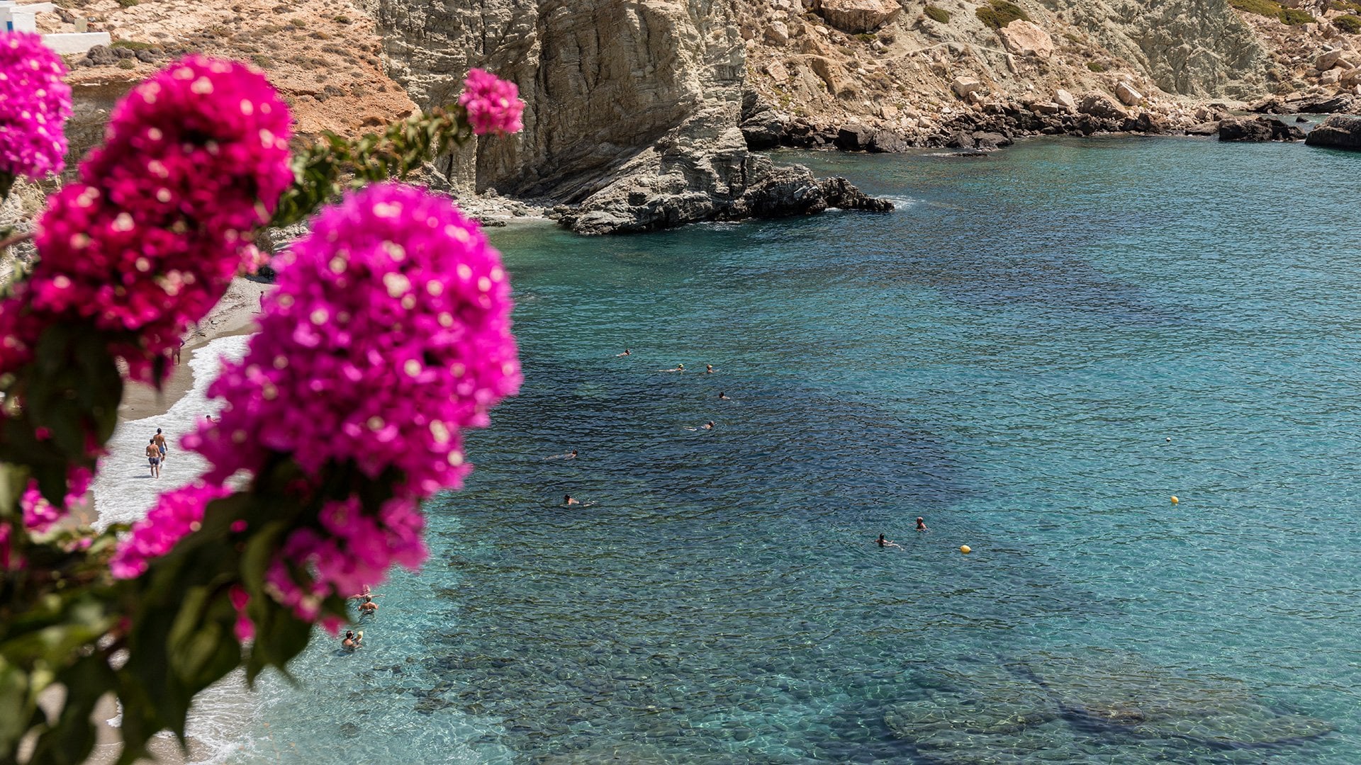 Gundari, Greece — Vibrant magenta bougainvillea framing turquoise Mediterranean cove with limestone cliffs and swimmers below.