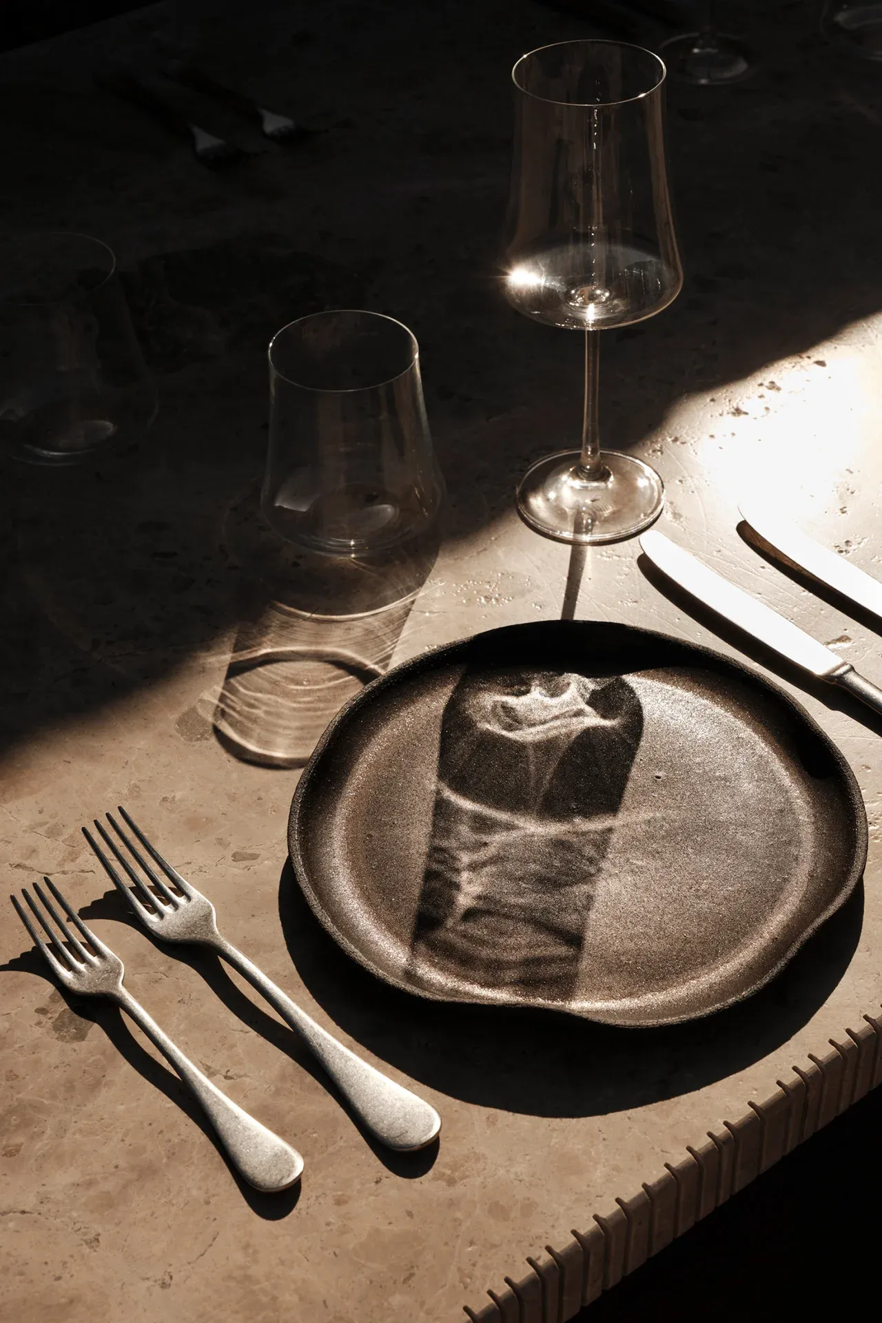 Gundari, Greece — Minimalist table setting with dark ceramic plate, folded napkin, forks, and wine glasses catching Mediterranean sunlight on stone surface.