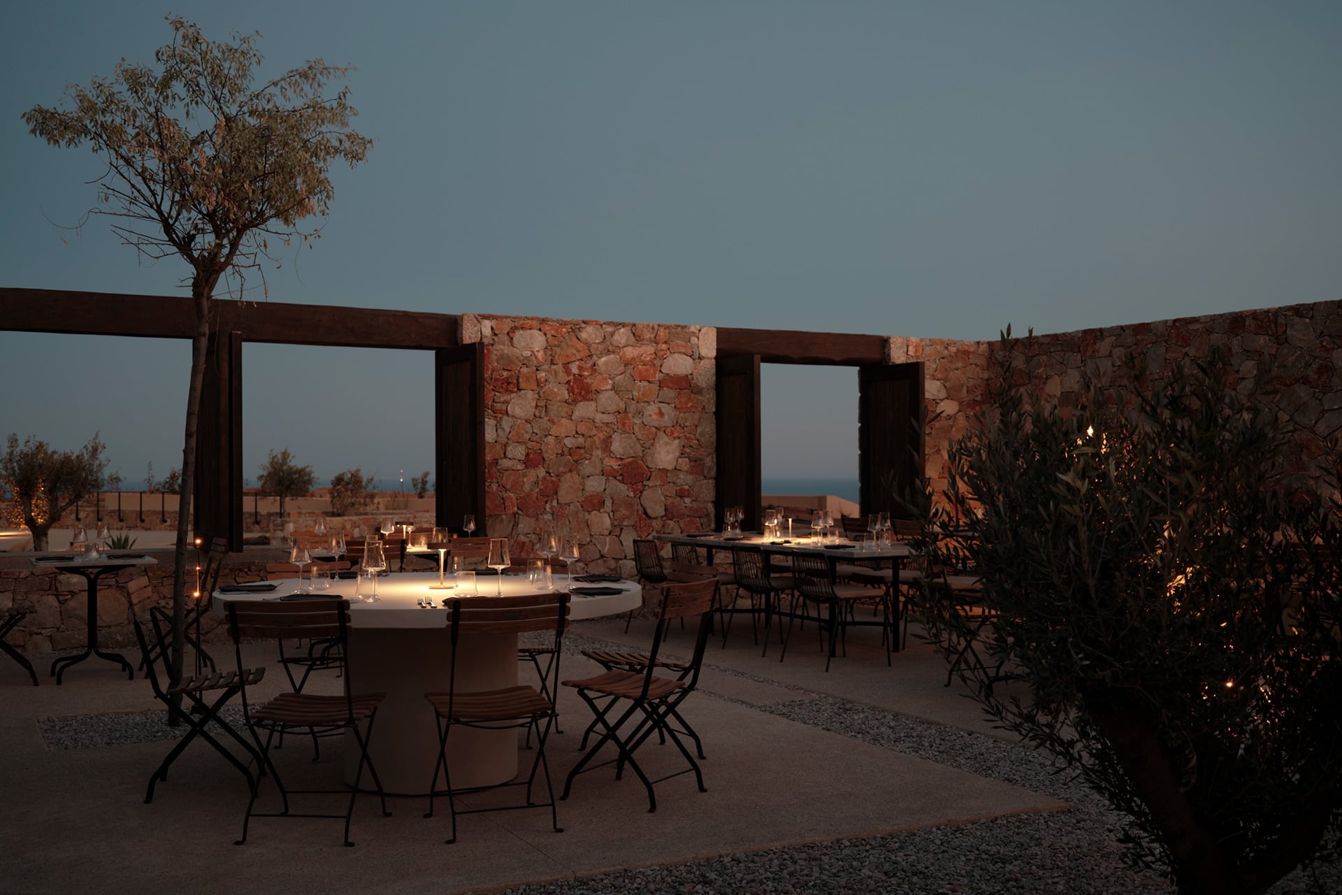 Orizon, Greece — Candlelit outdoor dining terrace at dusk with stone walls, metal pergolas, and sea views, modern hospitality design.