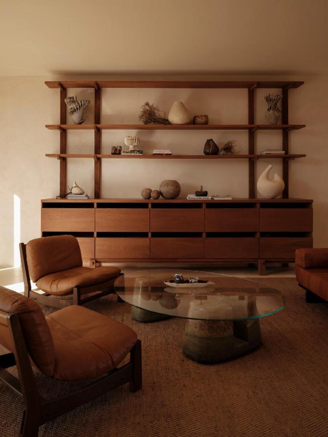 Gundari, Greece — Warm wooden shelving unit with sculptural ceramics, cognac leather seating, and circular glass table in minimalist interior.
