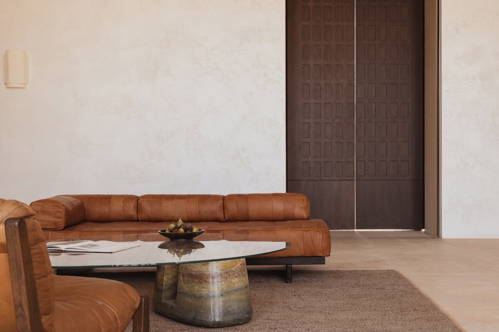 Gundari, Greece — Cognac leather sectional sofa with minimalist glass and stone coffee table in gallery space with textured white walls and dark wood doors.