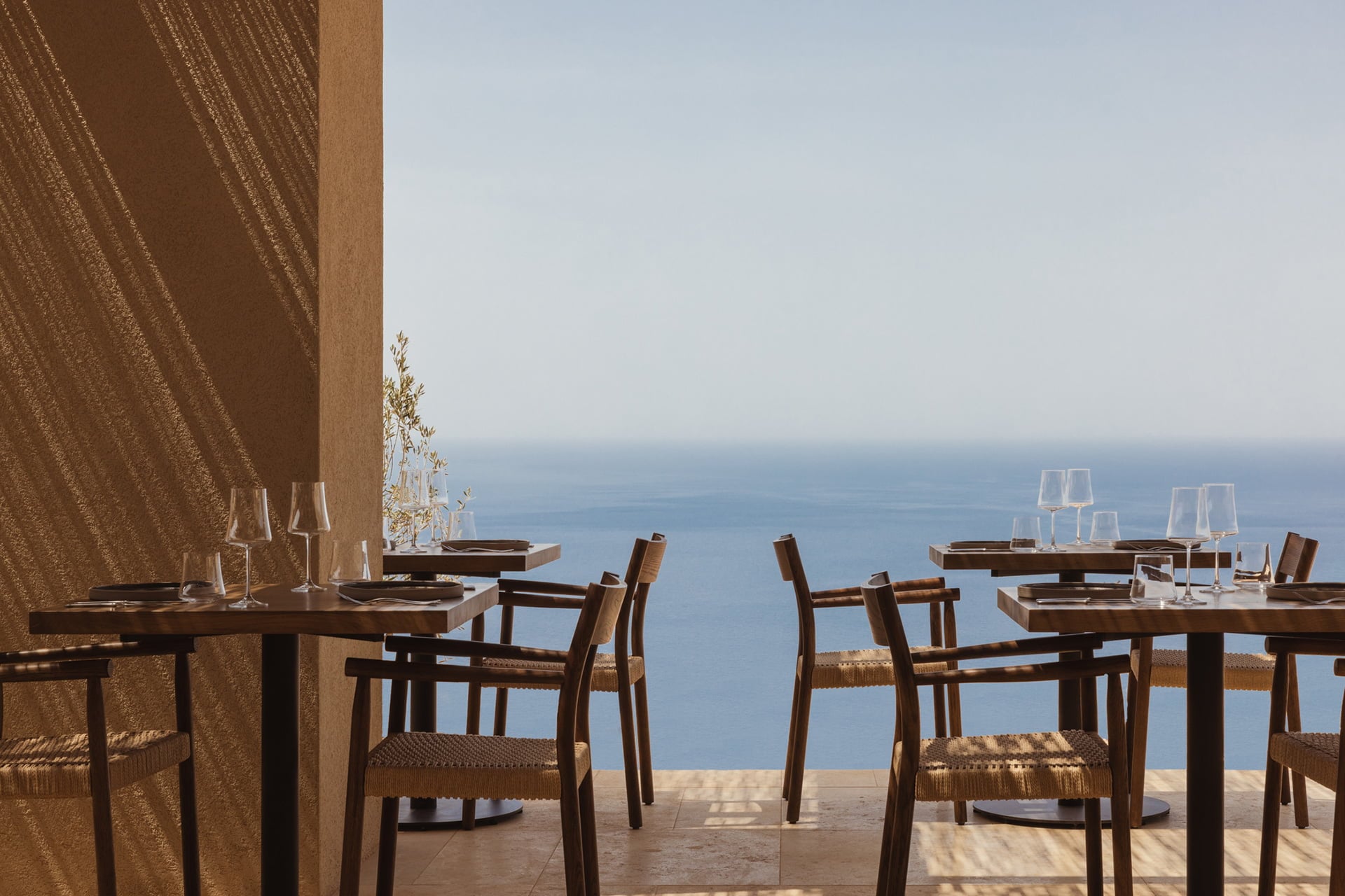 Gundari, Greece — Clifftop dining terrace with woven bronze shade, wooden chairs, and Aegean Sea views at sunset.