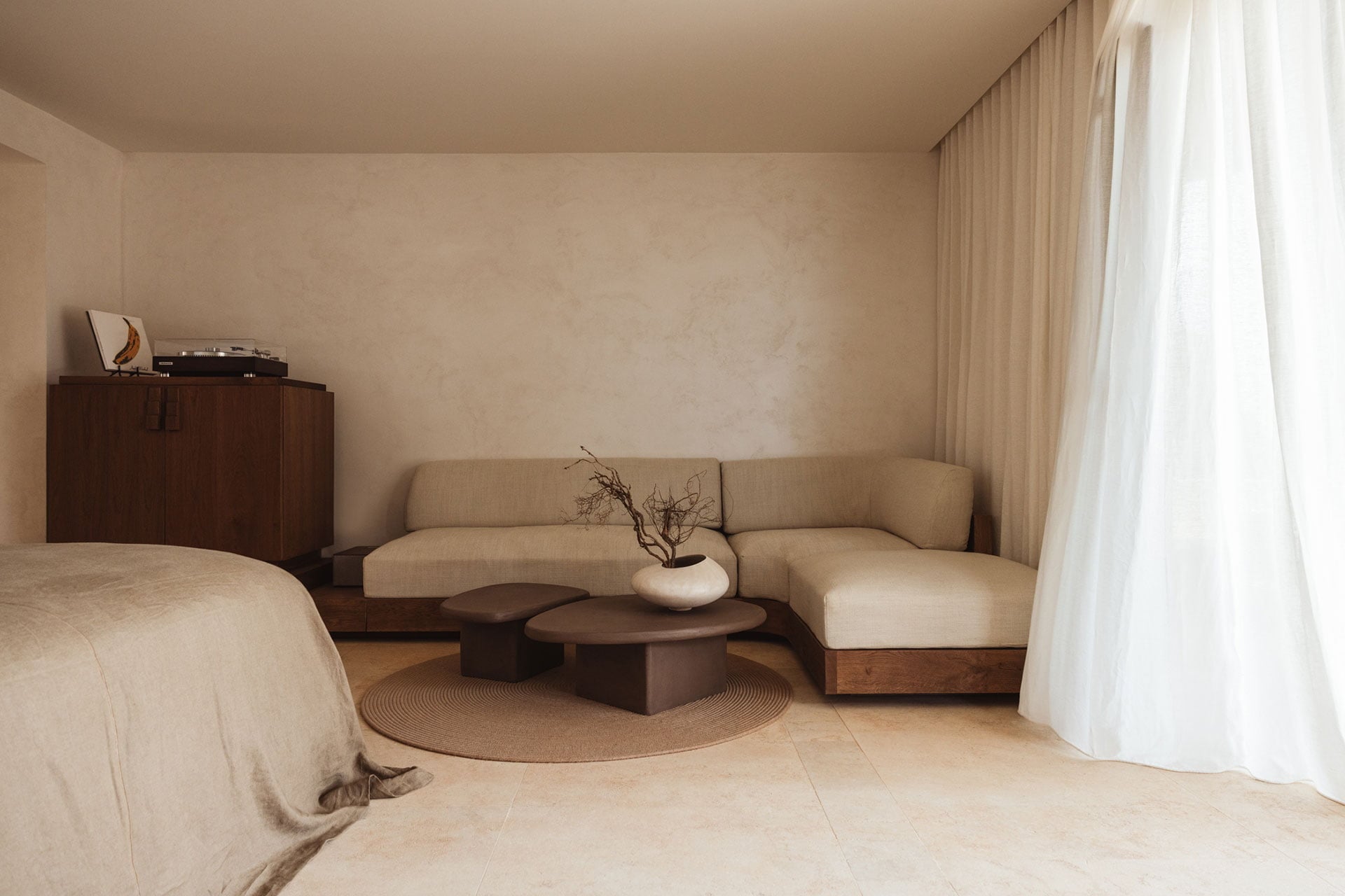 Gundari, Greece — Minimalist hotel suite with cream linen bed, curved beige sofa, dark wood furniture, circular rug, and soft natural light from sheer curtains.