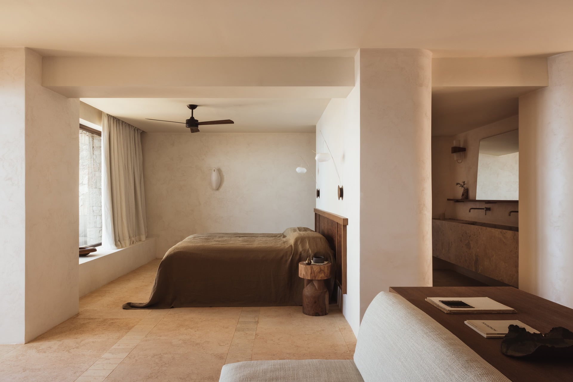 Gundari, Greece — Minimalist bedroom suite with terracotta flooring, warm plaster walls, wooden furnishings, and en-suite bathroom visible through open partition.