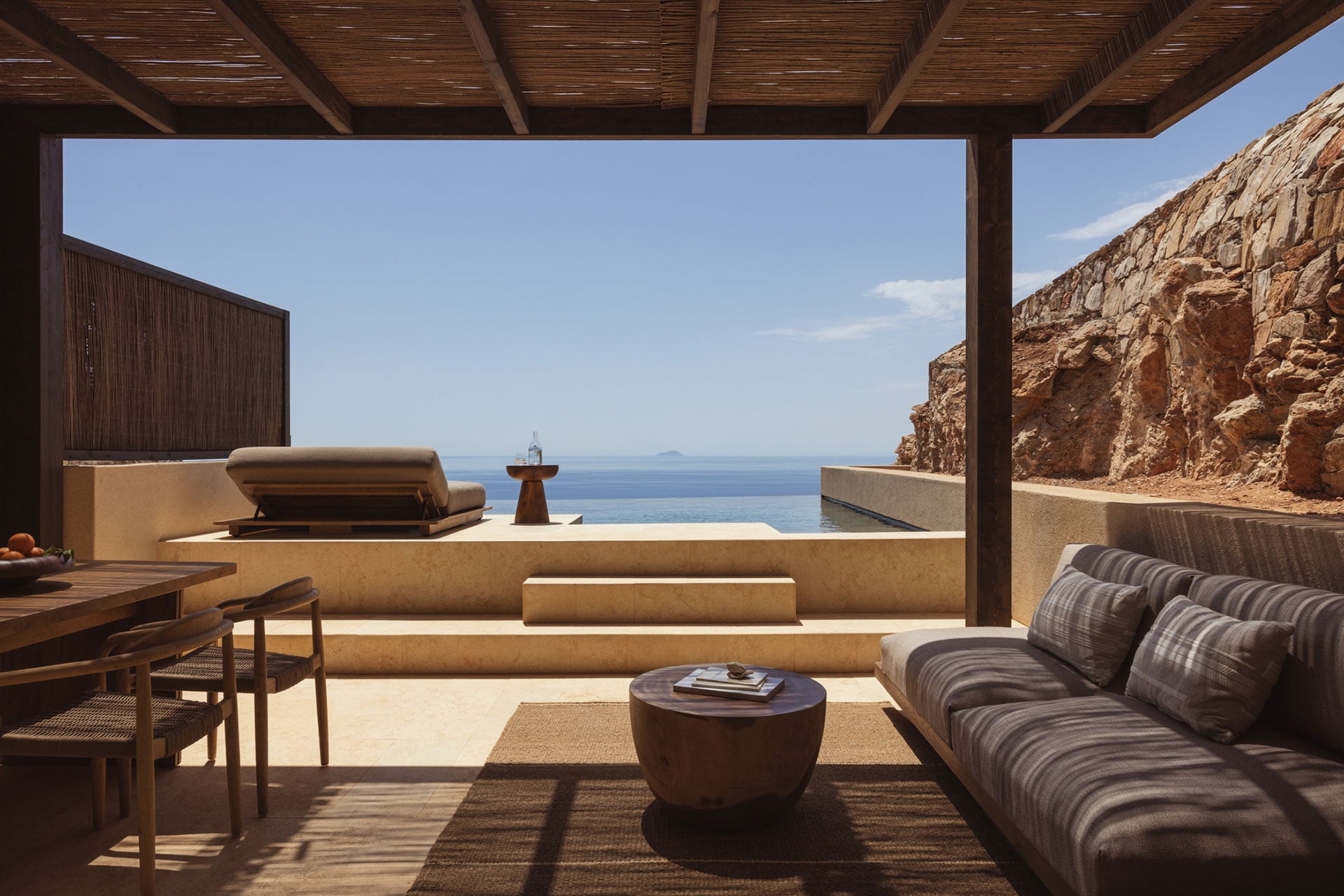 Gundari, Greece — Minimalist clifftop terrace with striped sofa, concrete bathtub, and Aegean Sea views framed by wooden pergola and raw stone walls.