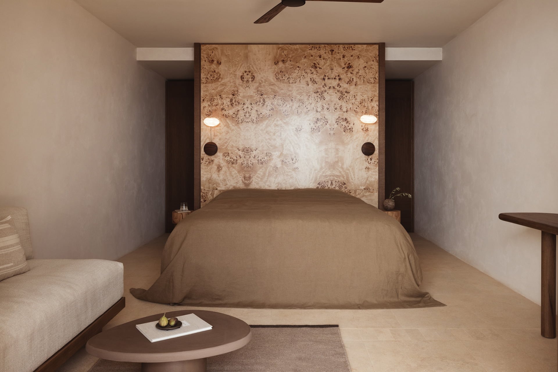 Gundari, Greece — Minimalist bedroom with burl wood headboard, warm taupe linens, recessed lighting, and curved accent table in neutral earth tones.