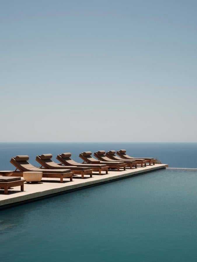 Gundari, Greece — Row of wooden loungers with cream cushions on pale stone terrace overlooking turquoise sea and horizon under clear sky.