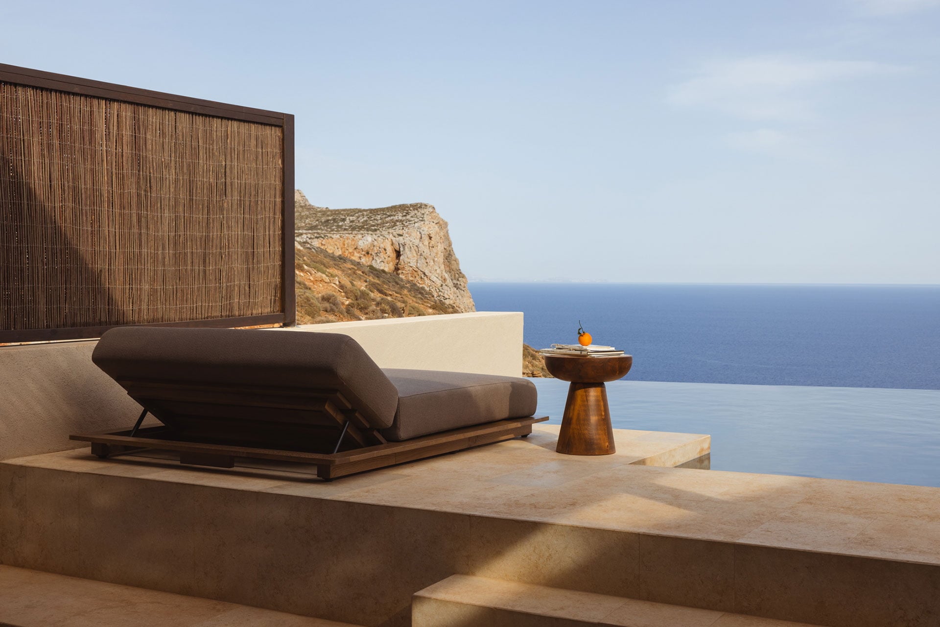 Gundari, Greece — Minimalist clifftop terrace with wooden lounge chair, stone platform, and Aegean Sea vista beyond rocky headland.