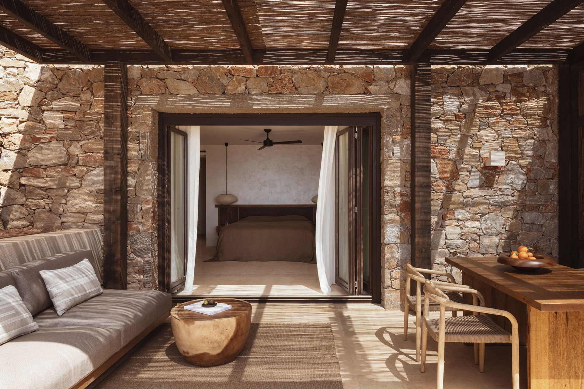 Gundari, Greece — Stone terrace with wooden pergola, daybed, and open bedroom doors overlooking minimalist interior with ceiling fan and natural light.