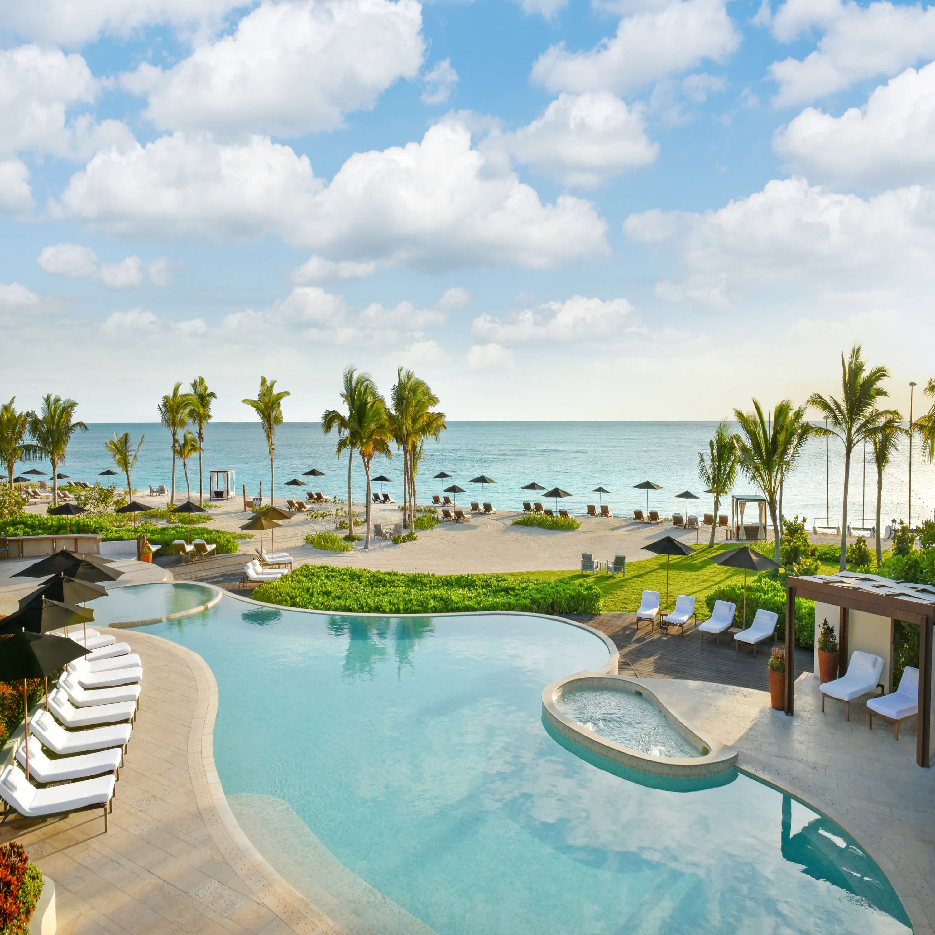 Rosewood Mayakoba, Mexico — Resort pool with curved edges, lounge chairs, spa, palm trees, and turquoise Caribbean Sea under clouded sky.