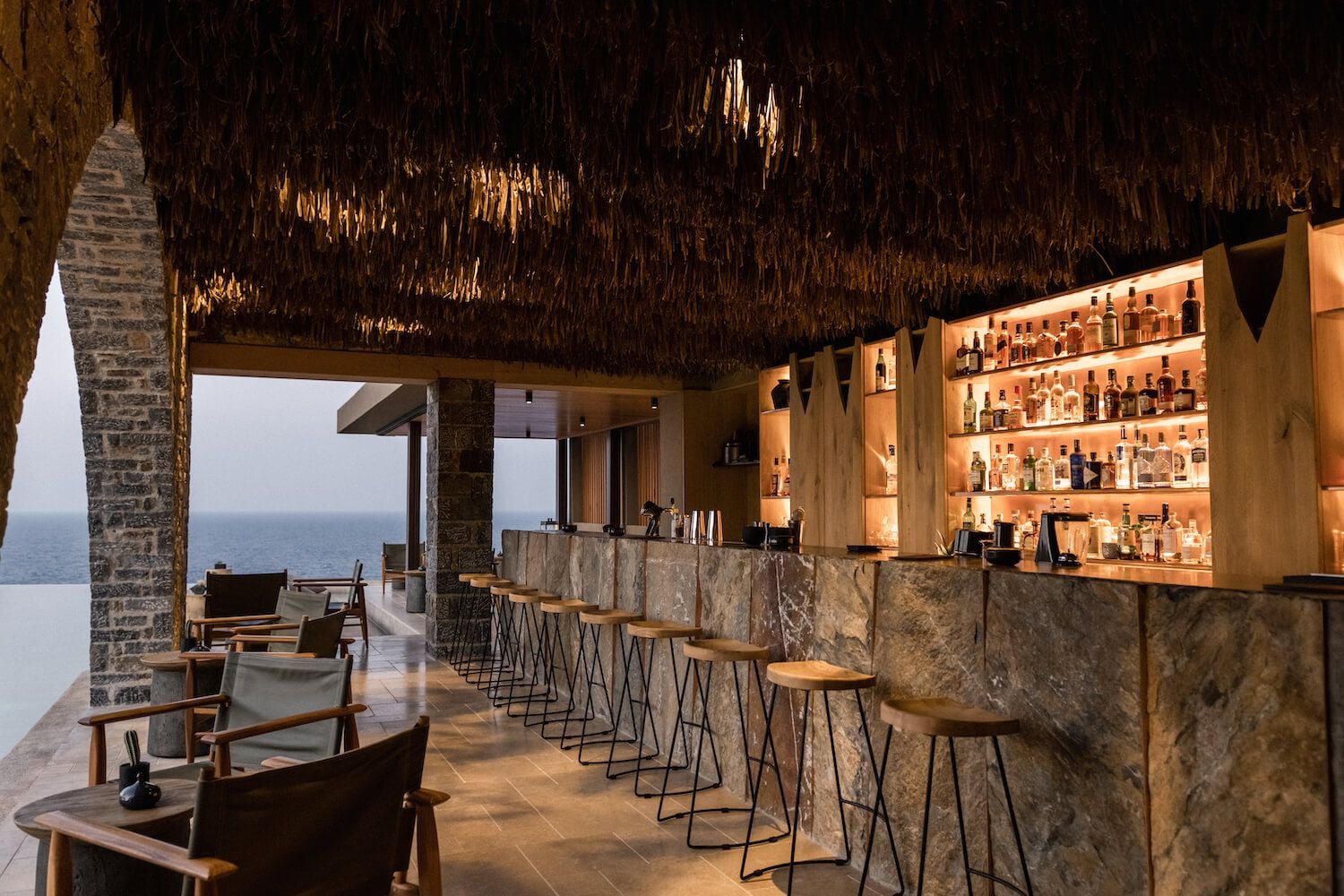 Pool Bar, Europe — Thatched ceiling bar overlooking Mediterranean sea with marble counter, wooden stools, backlit bottle shelves, and stone column detail.