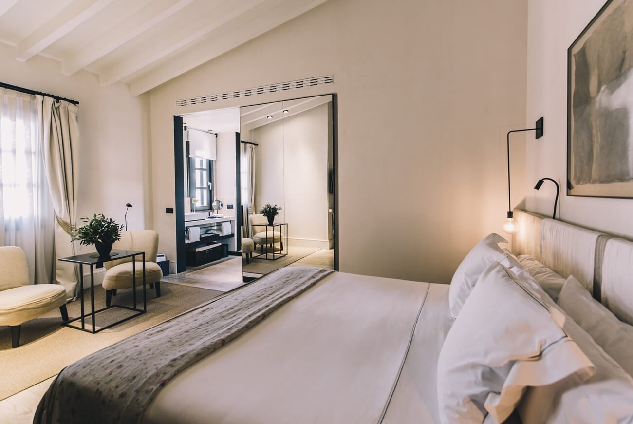 Sant Francesc Hotel Singular, Balearic Islands, Spain — Minimalist bedroom with neutral tones, exposed beams, and ensuite spa bathroom visible through open doors.