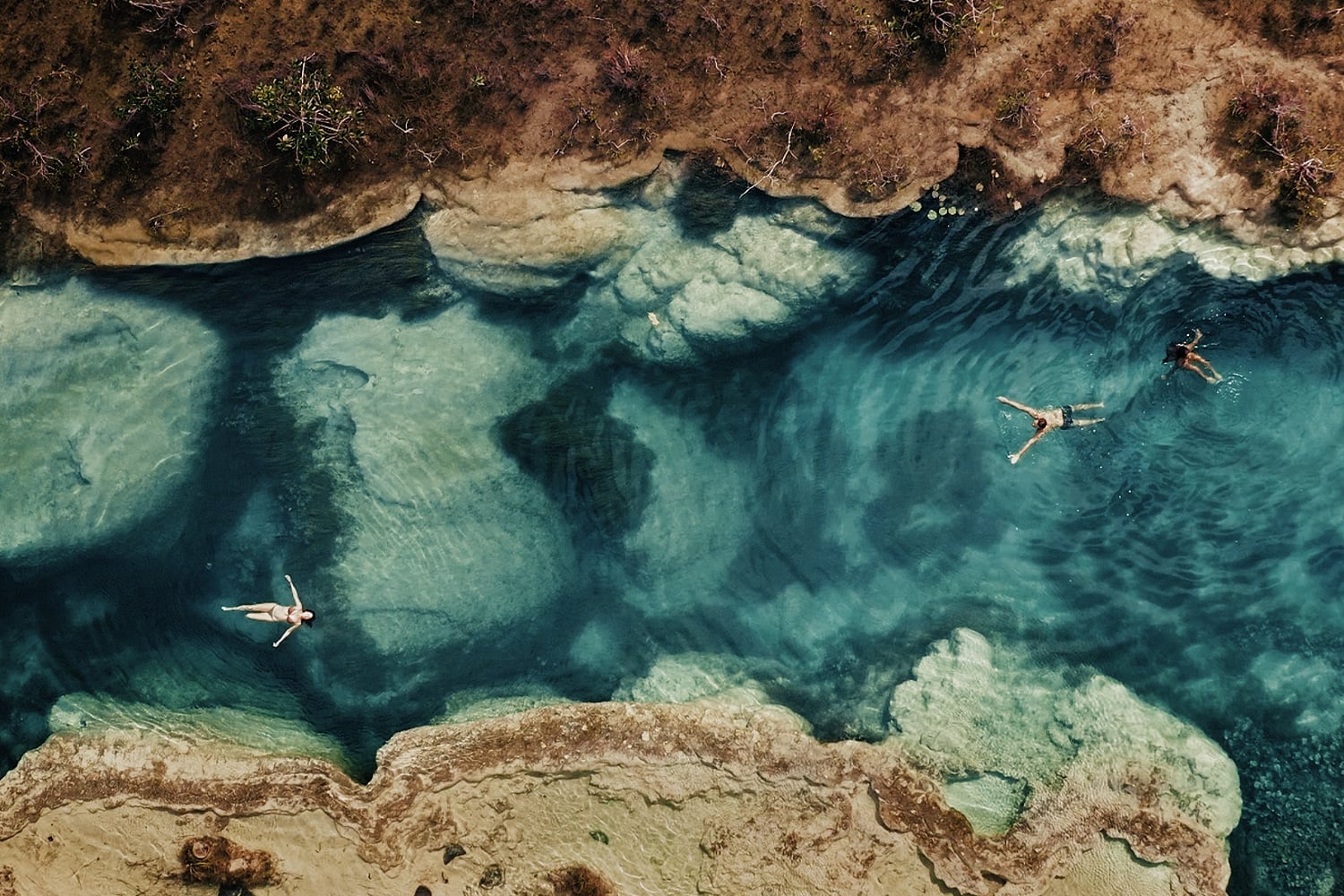 A New Sustainable Mexican Hotel — Aerial view of swimmers floating in crystalline turquoise water beside limestone cliffs and natural rocky formations.