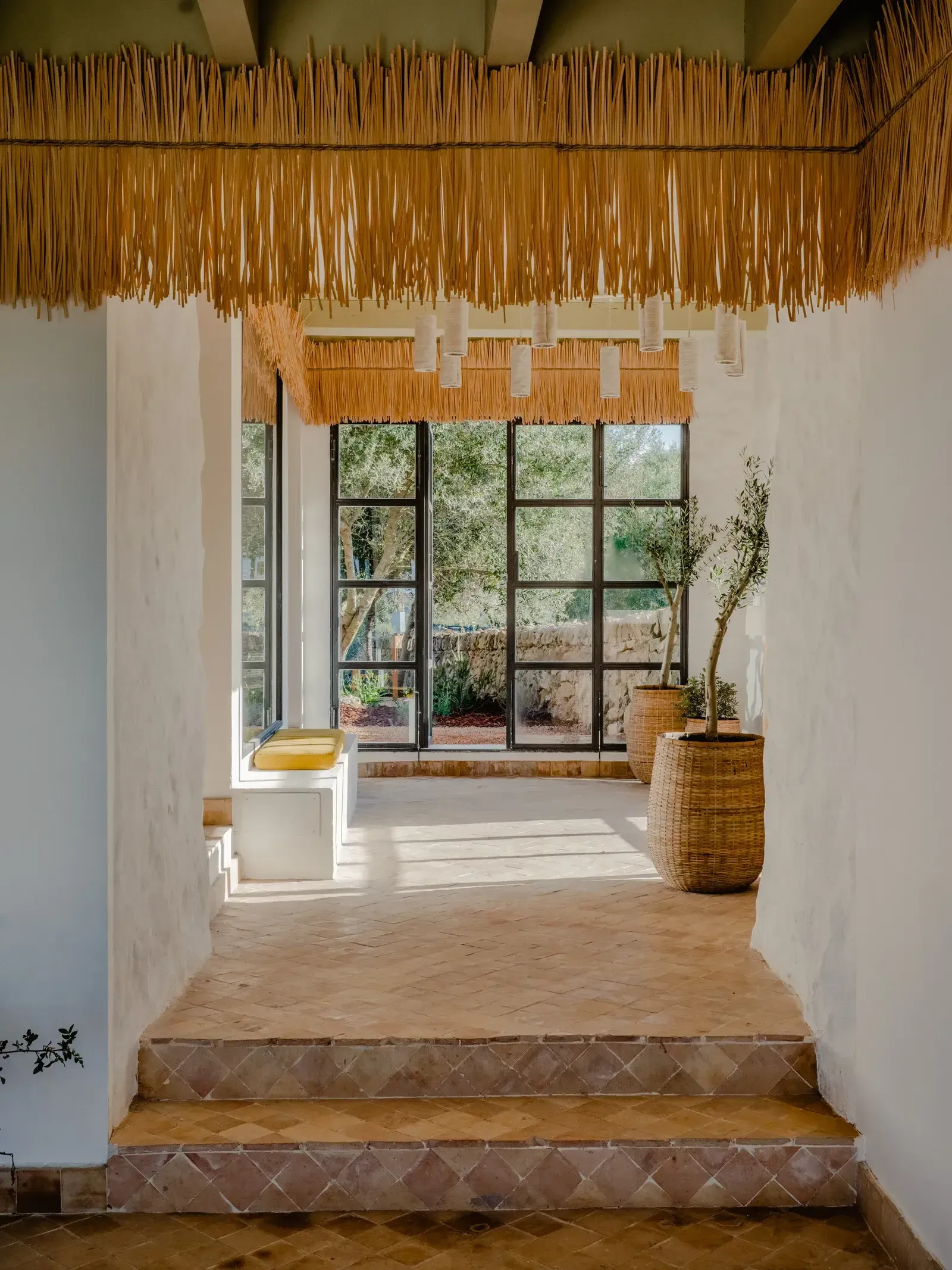 Amagatay, Balearic Islands — Minimalist corridor with thatched ceiling, terracotta tiles, woven planters, and gridded windows opening to Mediterranean landscape.
