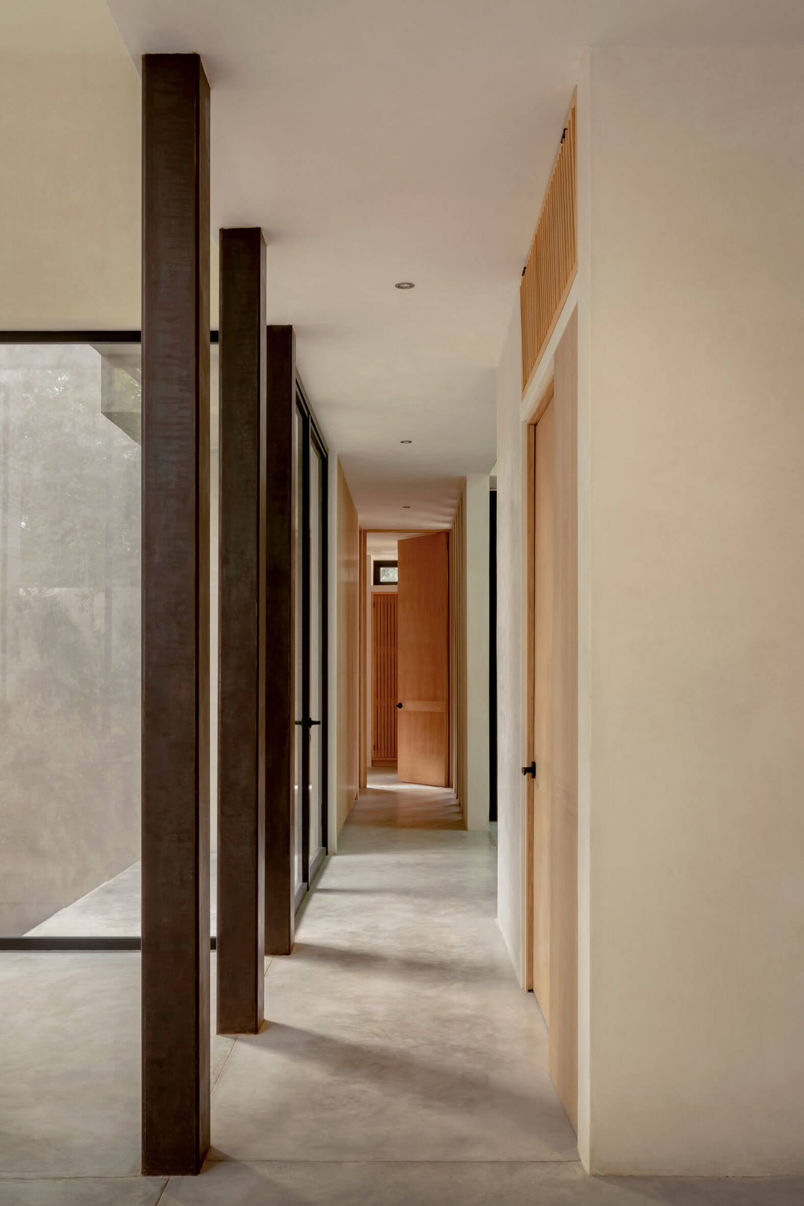 Villa Cava, Mexico — Minimalist hallway with dark wood columns, natural light, light stone flooring, and warm wood doors in modern luxury interior design.