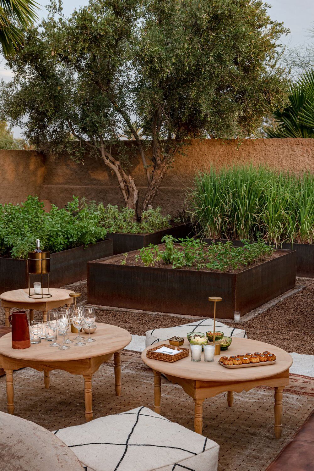 Villa D, Africa — Courtyard dining setup with wooden tables, metal-rimmed raised garden beds, olive tree, terracotta walls, and minimalist outdoor furniture.