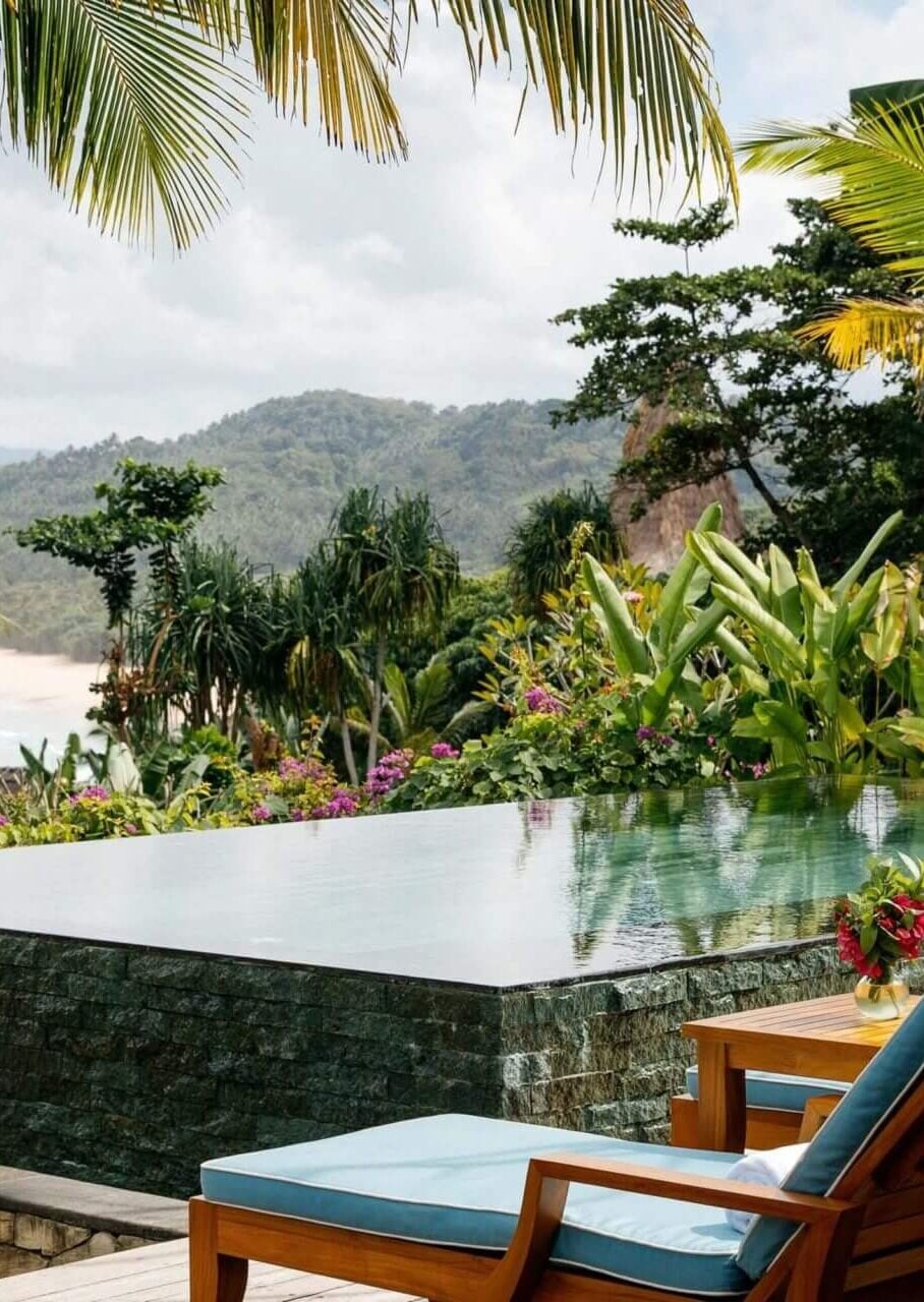 NIHI Sumba, Asia — Infinity pool with tropical gardens, palm fronds, flowering bougainvillea, and forested hillside beyond, stone terrace with teak loungers.