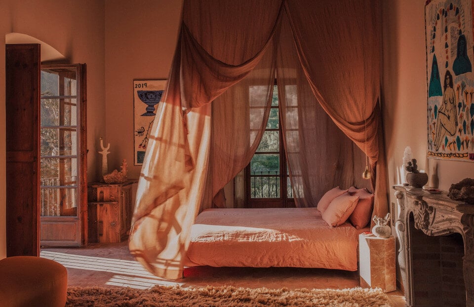 Hotel Corazón, Balearic Islands, Spain — Terracotta bedroom with sheer draped canopy, ochre sunlight across linen bedding, arched doorway, and decorative textile art on wall.