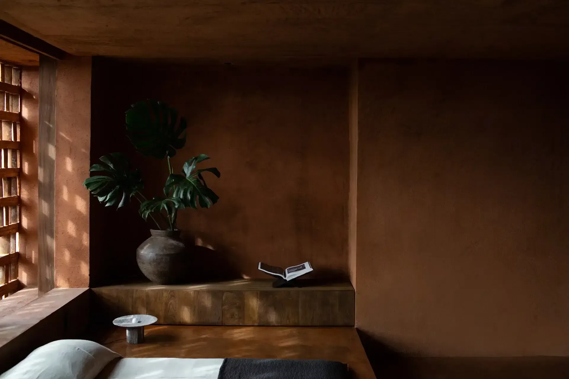 Further Hotel, Asia — Minimalist clay-toned alcove with wooden platform, potted monstera plant, open book, ceramic vessel, and warm natural light from latticed window.