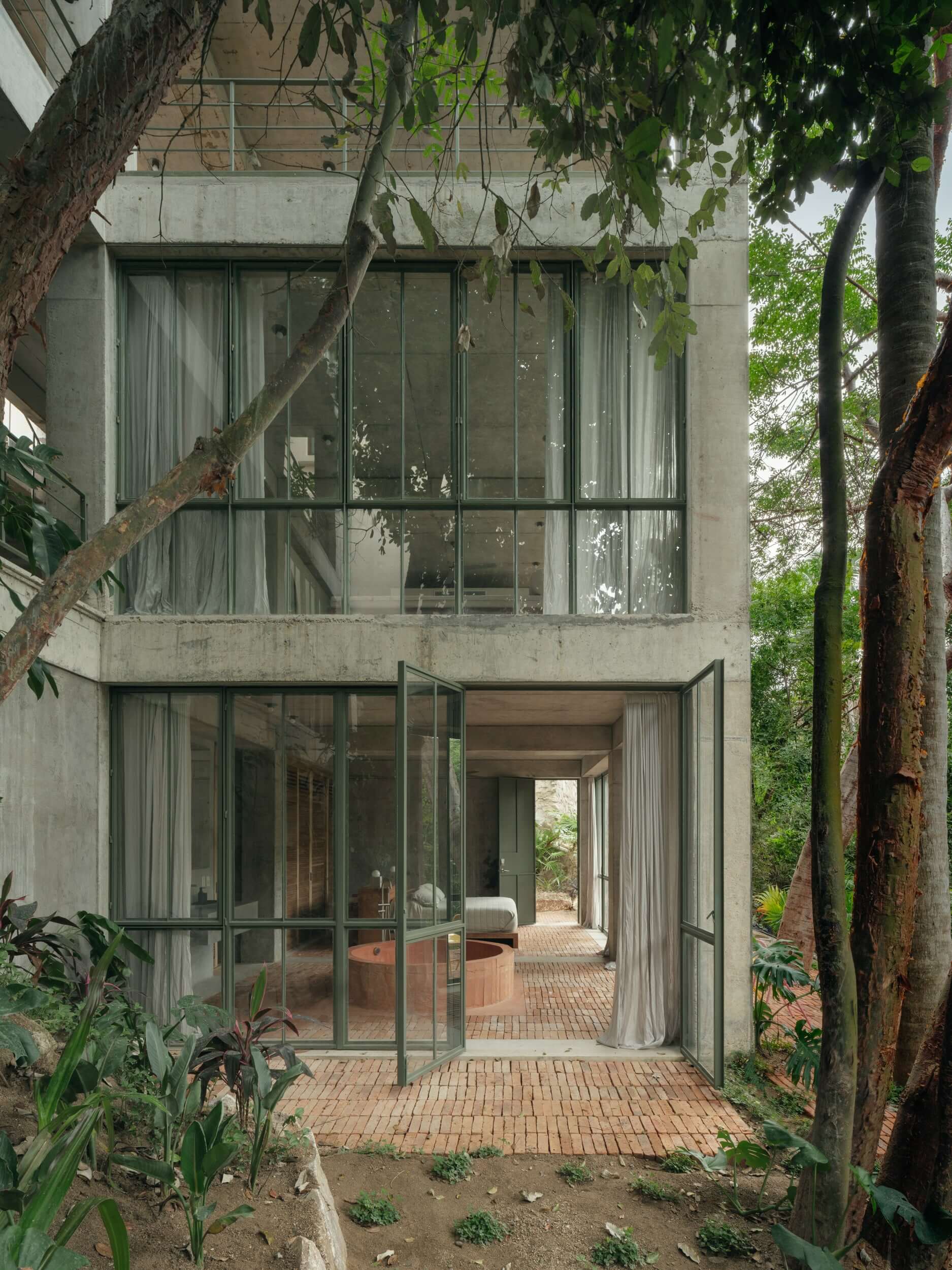 NICO, Mexico — Modernist concrete residence with floor-to-ceiling glazing, brick courtyard, and mature trees framing minimalist interiors and garden views.