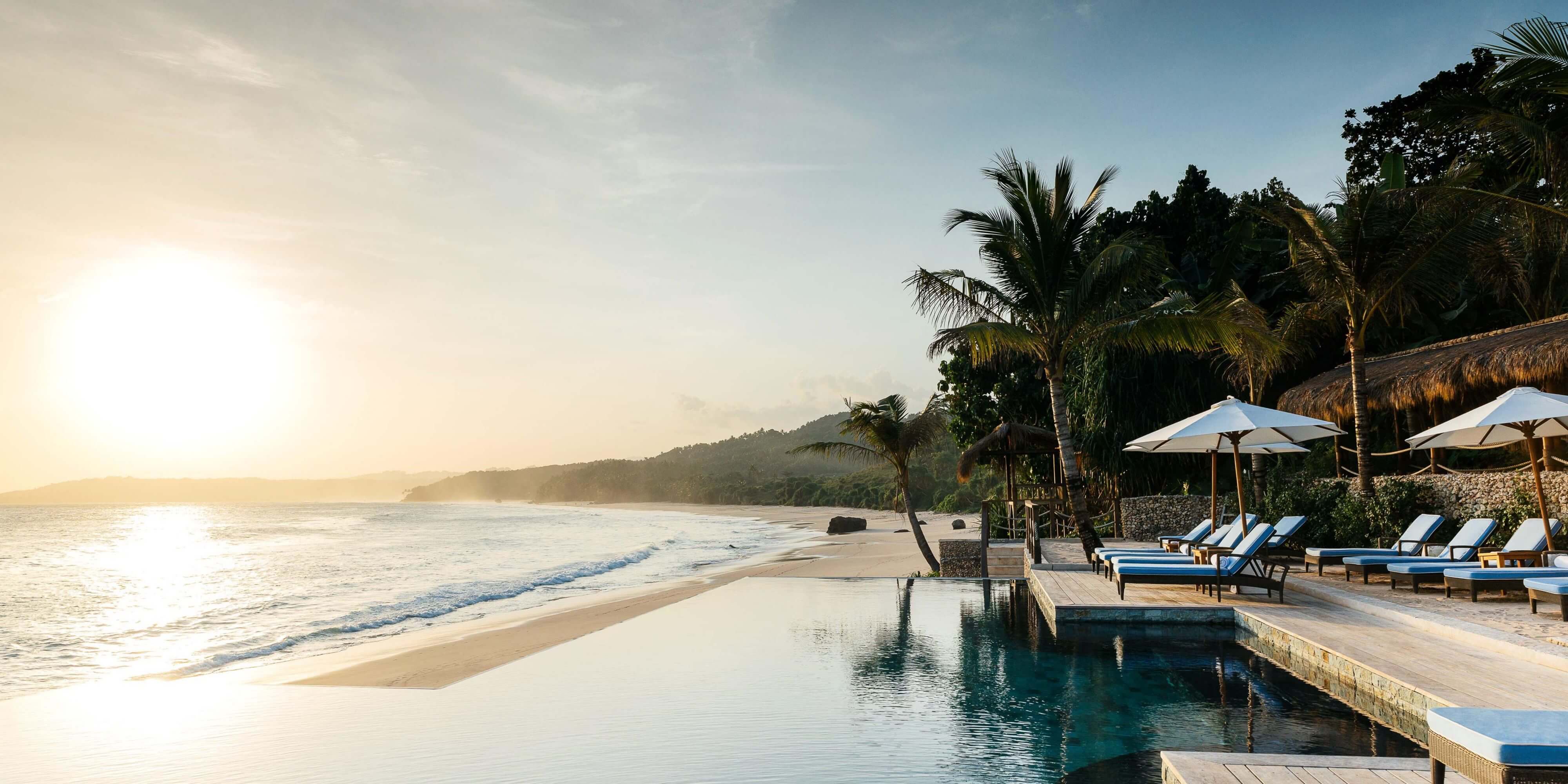NIHI Sumba, Asia — Infinity pool overlooking pristine beach at sunset, palm trees, blue loungers, thatched umbrellas, tropical coastline.