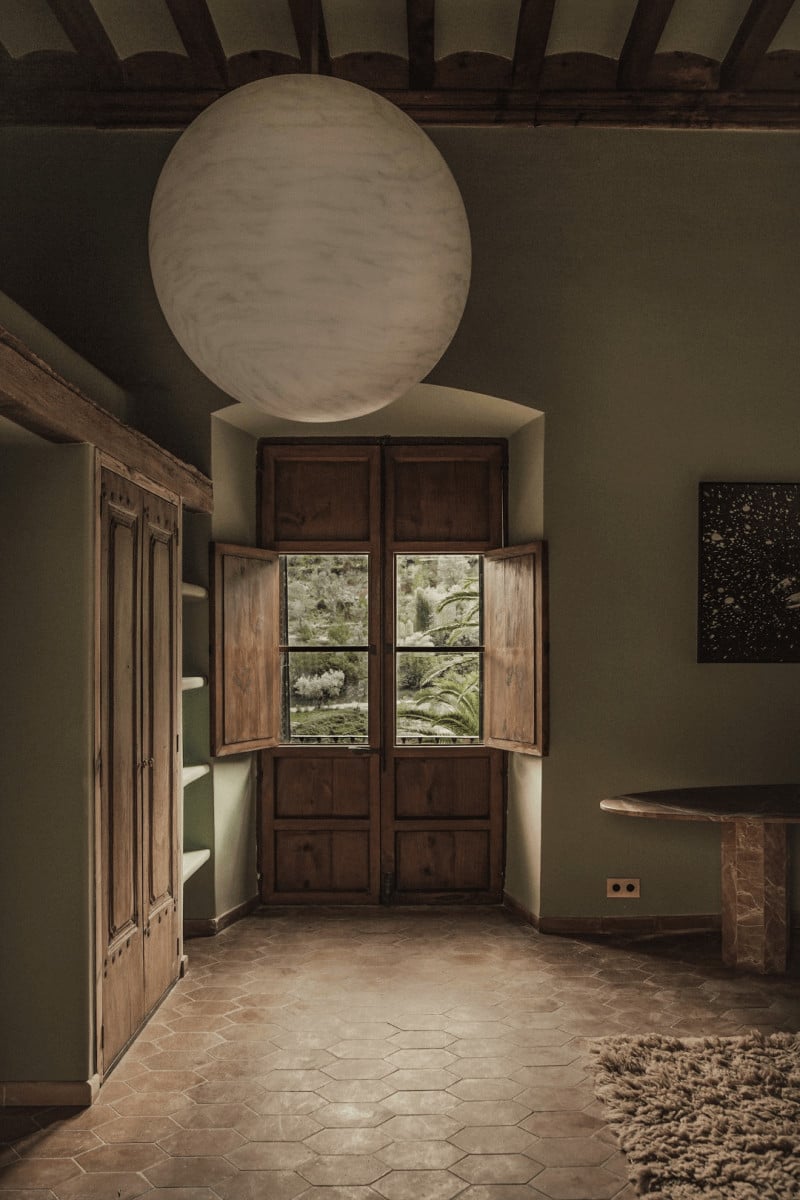 Hotel Corazón, Balearic Islands — Minimalist room with circular plaster skylight, dark wood doors opening to garden view, terracotta hexagonal floor tiles, exposed ceiling beams.