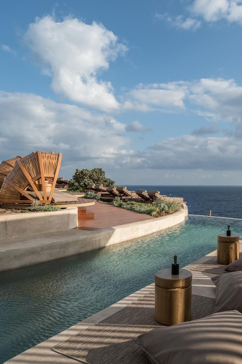 ACRO Suites, Europe — Infinity pool overlooking sea with wooden deck loungers, curved concrete architecture, and sculptural wooden shade structures against blue sky.