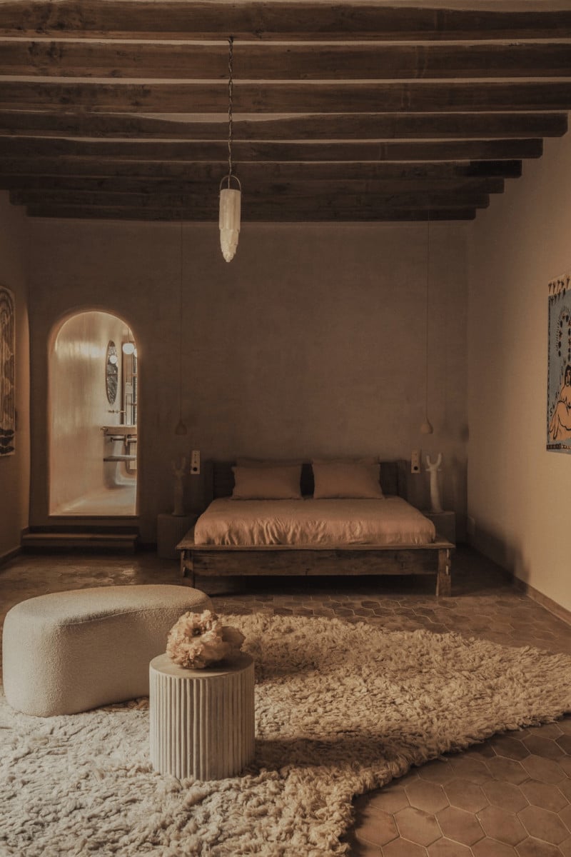 Hotel Corazón, Balearic Islands, Spain — Minimalist bedroom with exposed wooden beams, terracotta floors, linen furnishings, and layered natural fiber rugs in warm earth tones.
