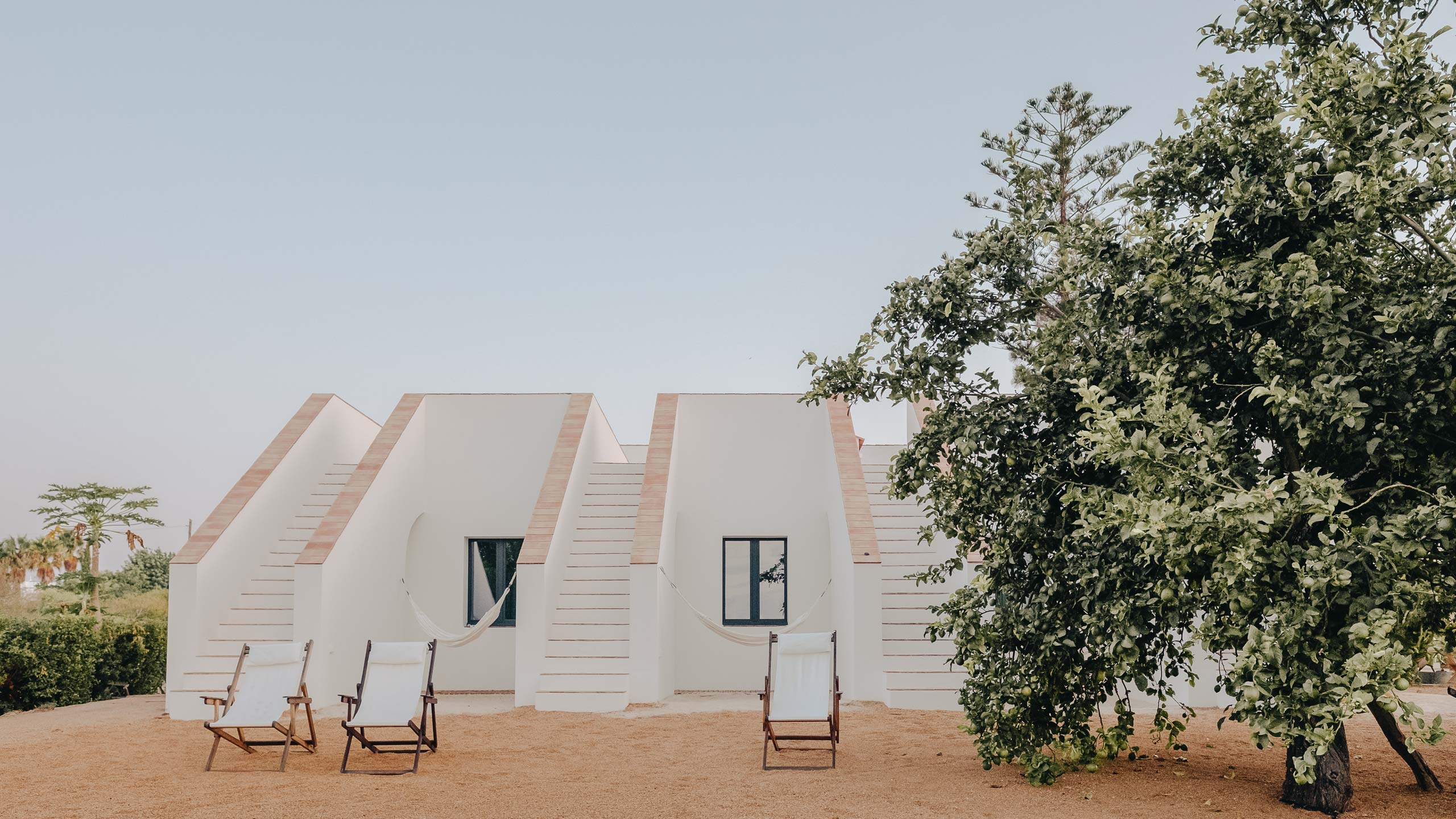 Casa Modesta, Europe — Minimalist white residential pavilions with terracotta trim, wooden deck chairs, and mature trees on sandy grounds under clear sky.