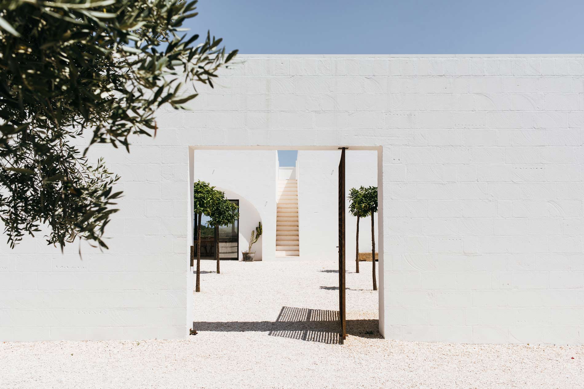 Masseria Moroseta, Europe — Minimalist courtyard with white-washed walls, terracotta staircase, timber pergola, potted topiaries, and gravel flooring in bright Mediterranean light.