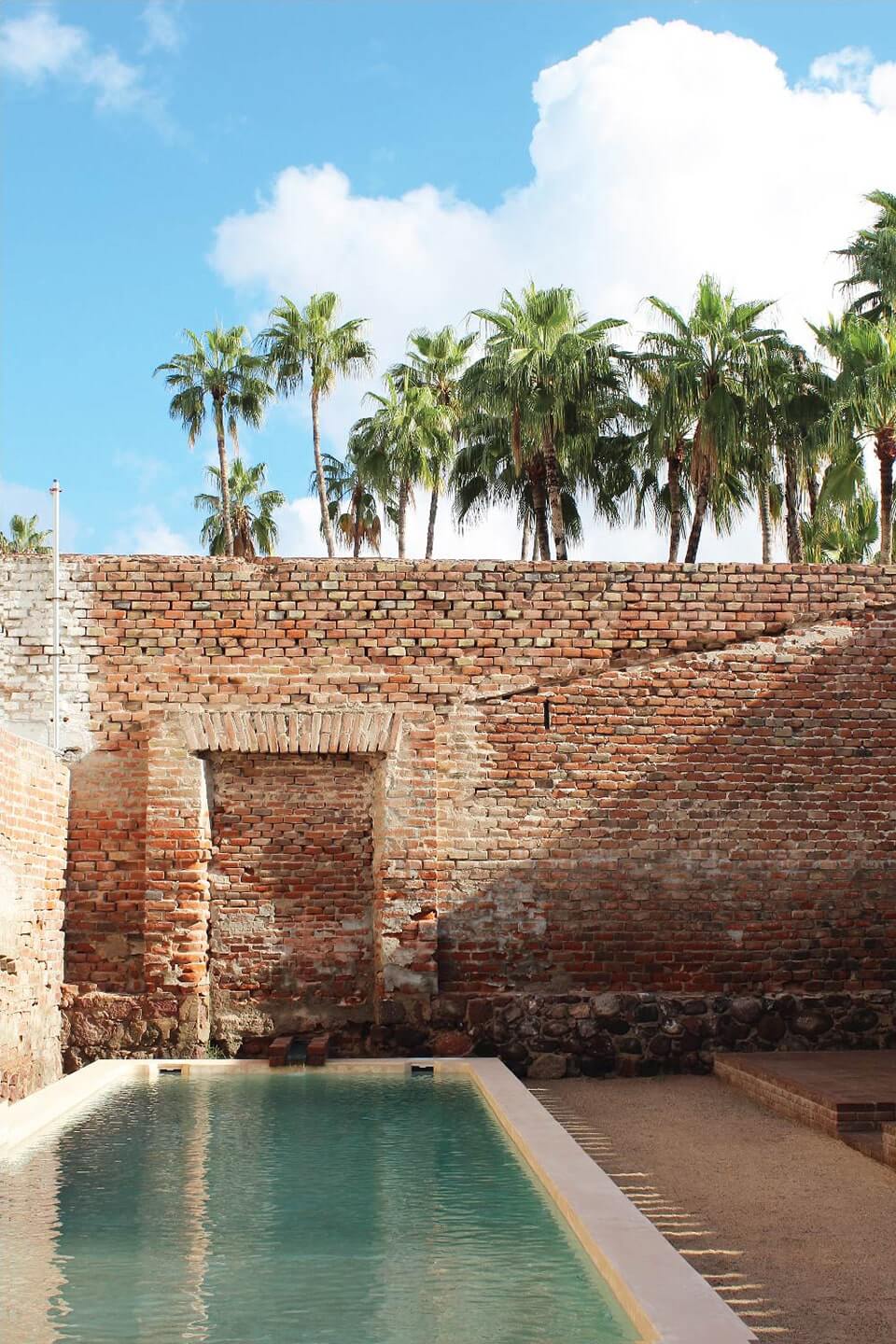 Baja Club, Mexico — Turquoise pool against weathered brick fort walls with arched doorway and palm trees under blue sky.