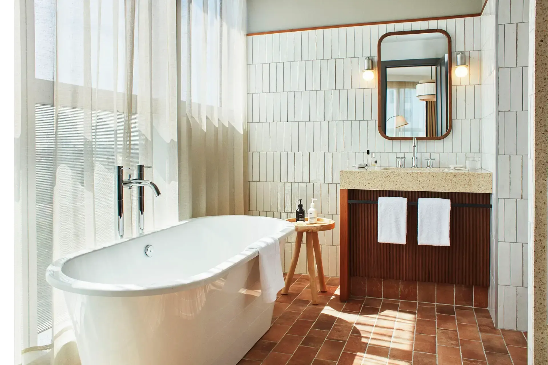 The Hoxton Poblenou, Barcelona, Spain — Spa bathroom with freestanding soaker tub, white subway tile, terrazzo vanity, wooden cabinetry, bronze mirror, and warm natural light from large windows.