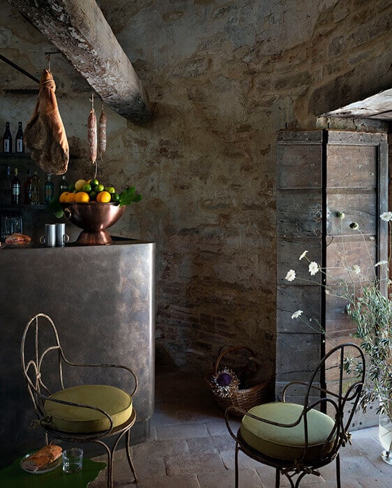 Castello di Reschio, Europe — Rustic stone kitchen with aged concrete walls, iron chairs, copper vessels, hanging cured meats, and weathered metal storage cabinets.