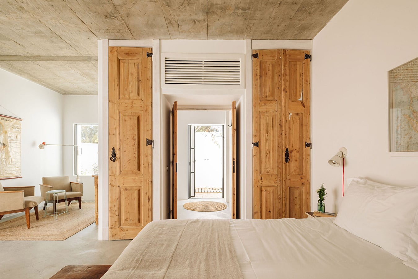 Hospedaria, Europe — Minimalist bedroom with wooden doors, concrete ceiling, natural light, and open terrace access, featuring mid-century furniture and neutral palette.