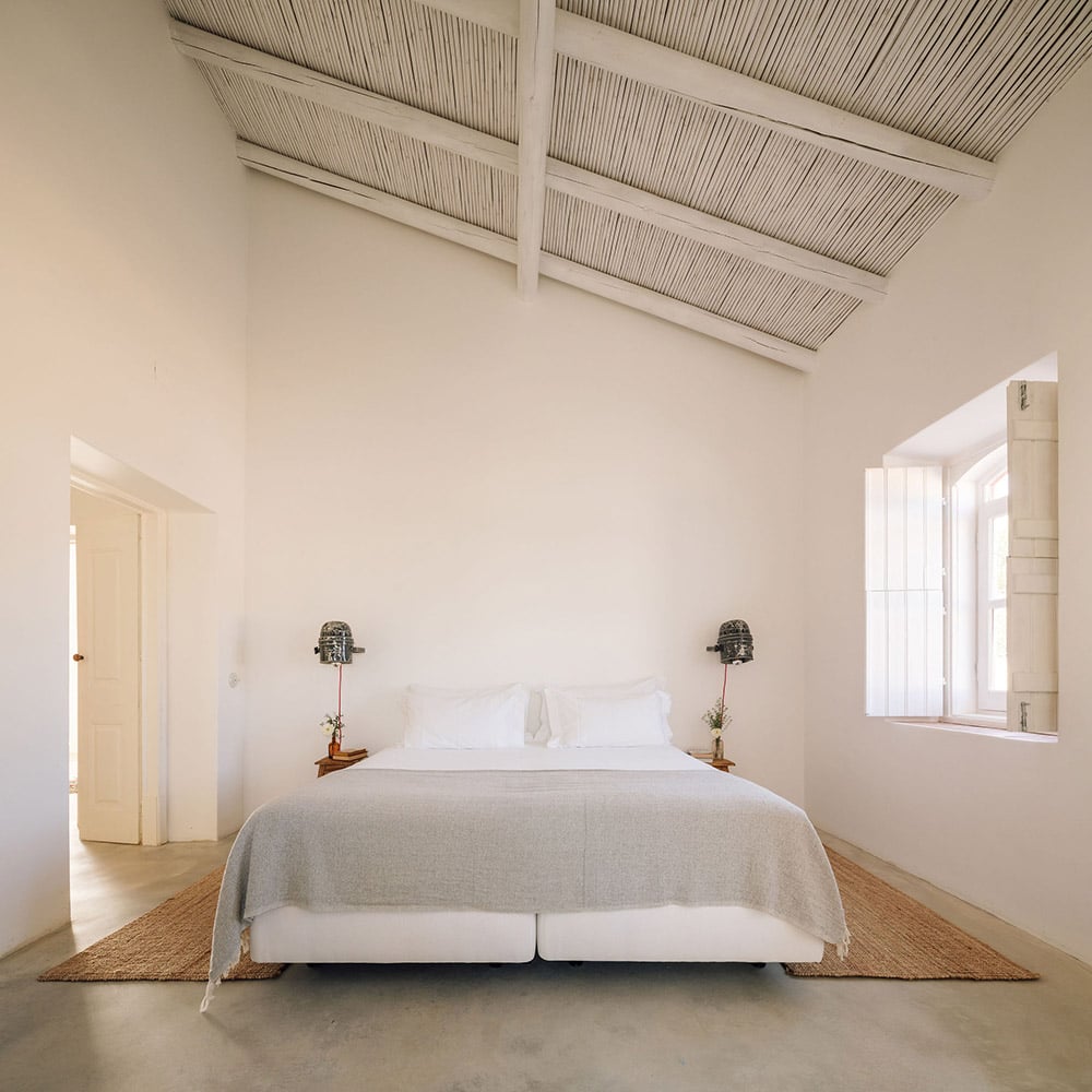 Hospedaria, Europe — Minimalist bedroom with skylight, concrete floors, linen bedding, wooden nightstands, and natural light from large windows.