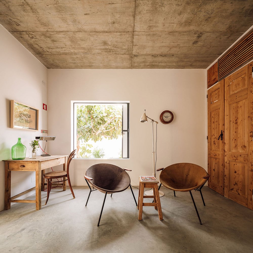 Hospedaria, Europe — Minimalist hotel room with exposed concrete ceiling, wooden furniture, mid-century modern chairs, natural light, and rustic-industrial aesthetic.
