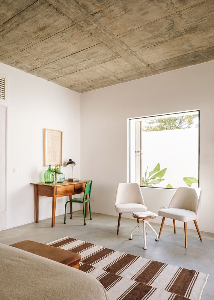 Hospedaria, Europe — Minimalist hotel room with exposed wooden ceiling, cream walls, mid-century furniture, concrete floors, and bright window overlooking greenery.