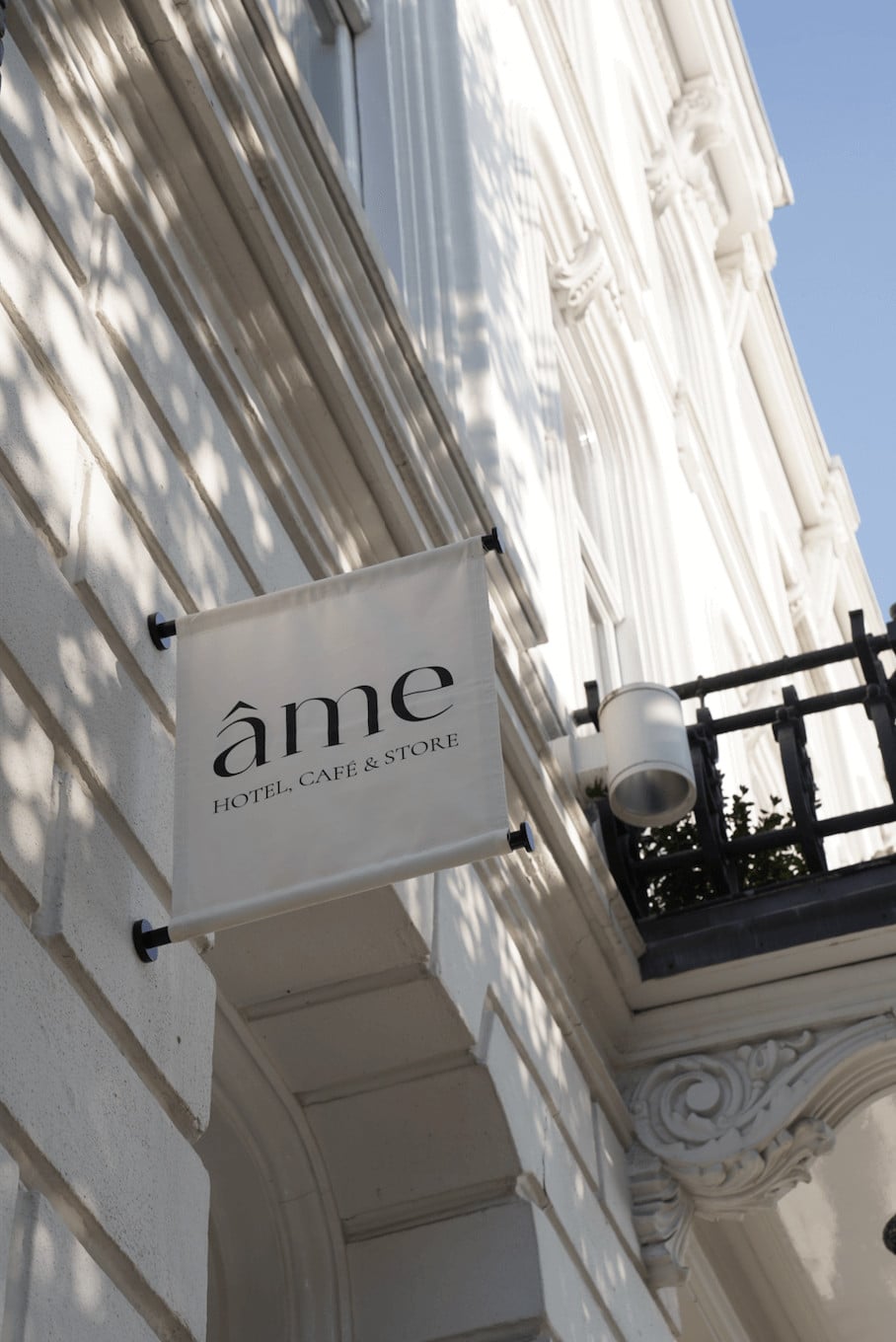 Hotel âme, Europe — White neoclassical facade with vertical fluting and ornamental details, minimalist signage with black brackets casting sharp shadows in bright daylight.