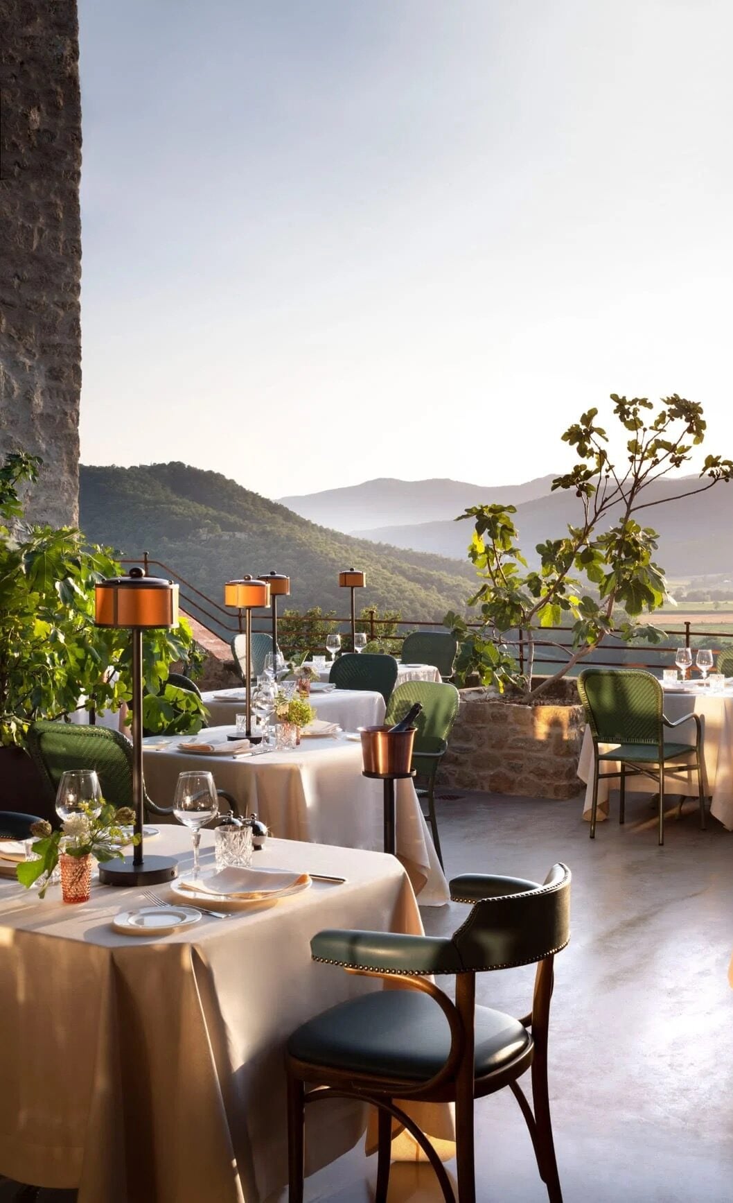 Castello di Reschio, Umbria, Italy — Hilltop terrace dining with copper heaters, linen-draped tables, green leather chairs, and panoramic valley views at sunset.