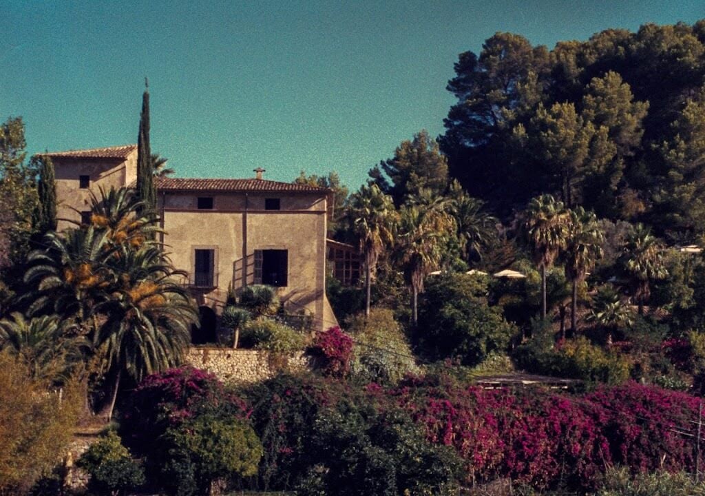 Hotel Corazón, Balearic Islands, Spain — Ochre stone villa with cypress and palm trees, magenta bougainvillea, Mediterranean garden landscape, clear turquoise sky.